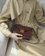 Model sitting on a couch wearing a beige trench dress holding a dark brown leather envelope flap shoulder bag.