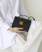 Model in a white outfit holding a black structured leather envelope bag with a gold lock closure on her lap.