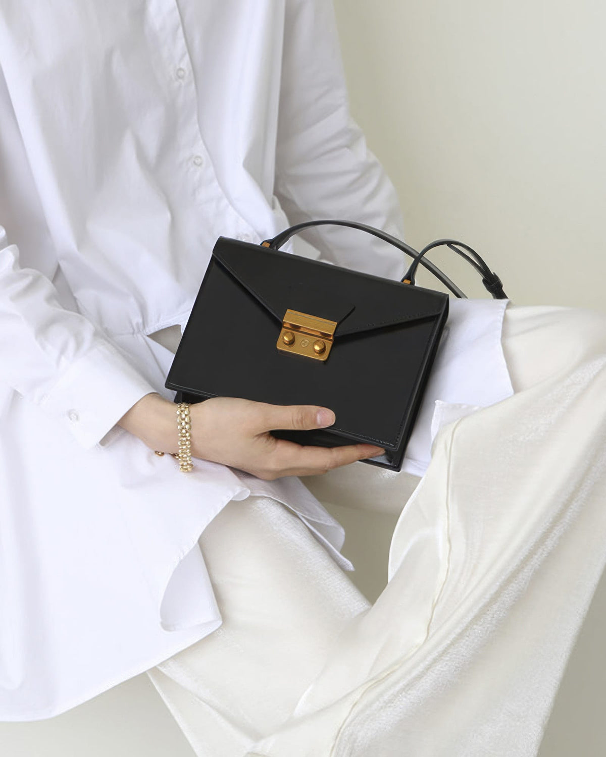 Model in a white outfit holding a black structured leather envelope bag with a gold lock closure on her lap.