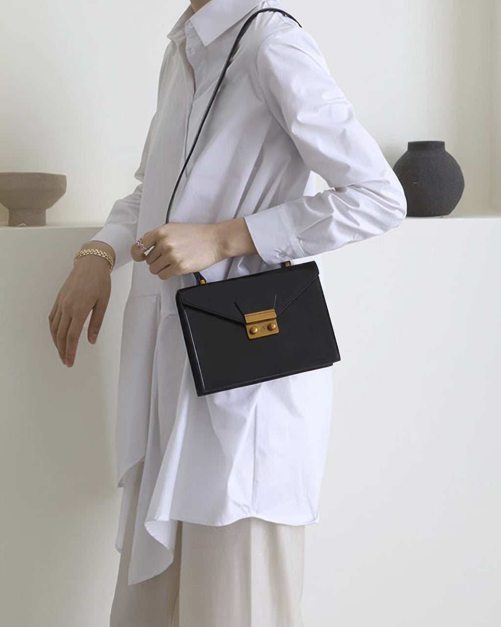 Side view of a model in a white long-sleeve shirt wearing a black leather envelope crossbody handbag with gold hardware.