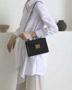 Side view of a model in a white long-sleeve shirt wearing a black leather envelope crossbody handbag with gold hardware.
