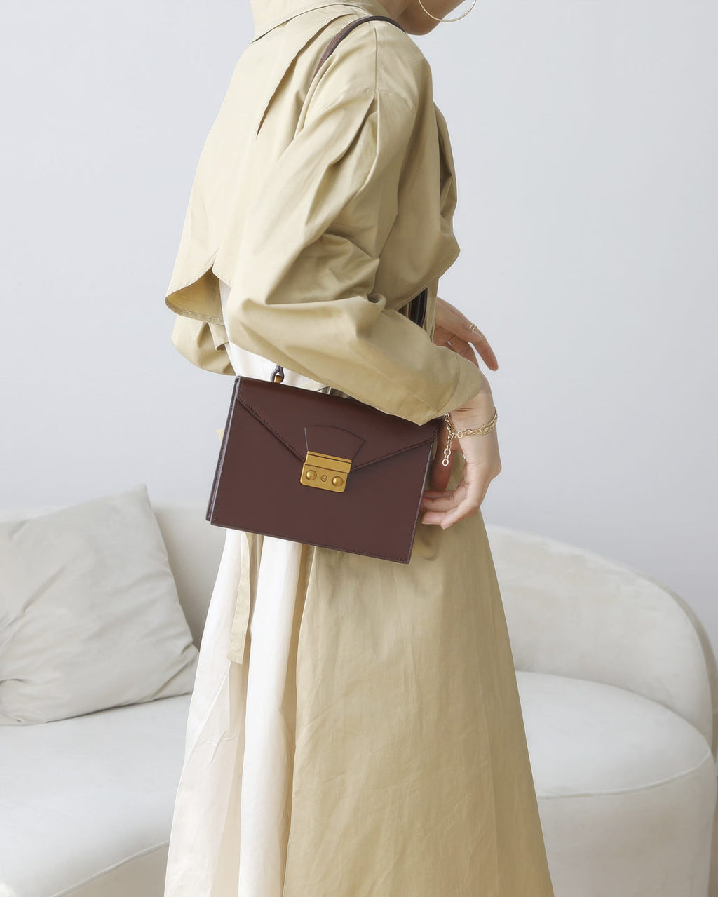 Model in a beige trench coat carrying a dark brown leather envelope flap handbag over her shoulder.