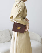 Model in a beige trench coat carrying a dark brown leather envelope flap handbag over her shoulder.