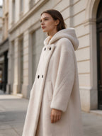 Low-angle side view of a model in a warm white hooded double-breasted shearling long coat walking outside.