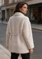  Back profile view of a woman wearing an off-white diamond lattice shearling wool coat with a drawstring waist outdoors.