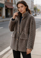 Side angle of a model styling a taupe brown quilted shearling wool coat with gold buttons and front flap pockets outdoors.