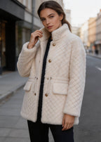 Side angle of a woman styling an off-white quilted shearling wool coat with a high collar and decorative buttons outdoors.