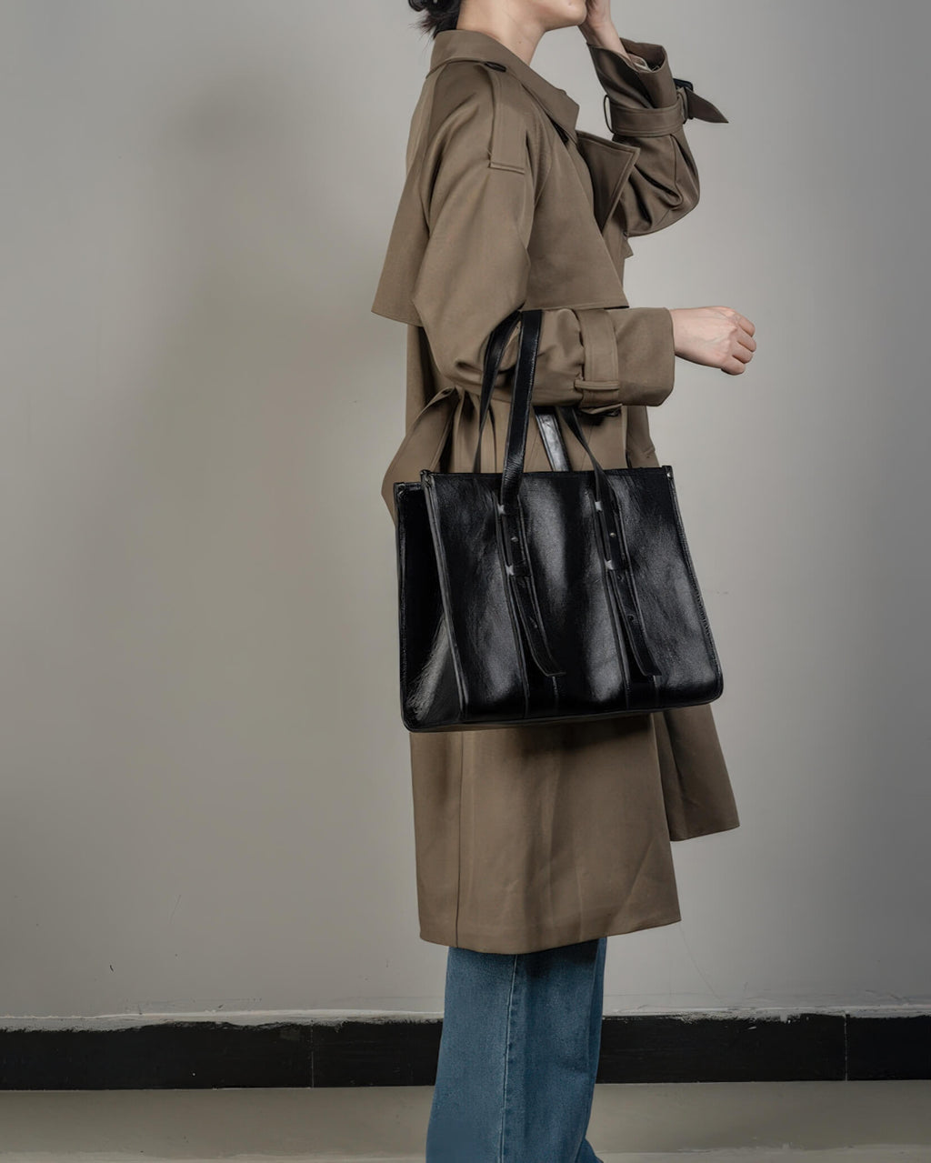 Fashion lifestyle shot of a woman carrying a black genuine leather tote bag on her shoulder while wearing a classic trench coat.