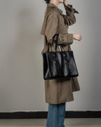 Fashion lifestyle shot of a woman carrying a black genuine leather tote bag on her shoulder while wearing a classic trench coat.