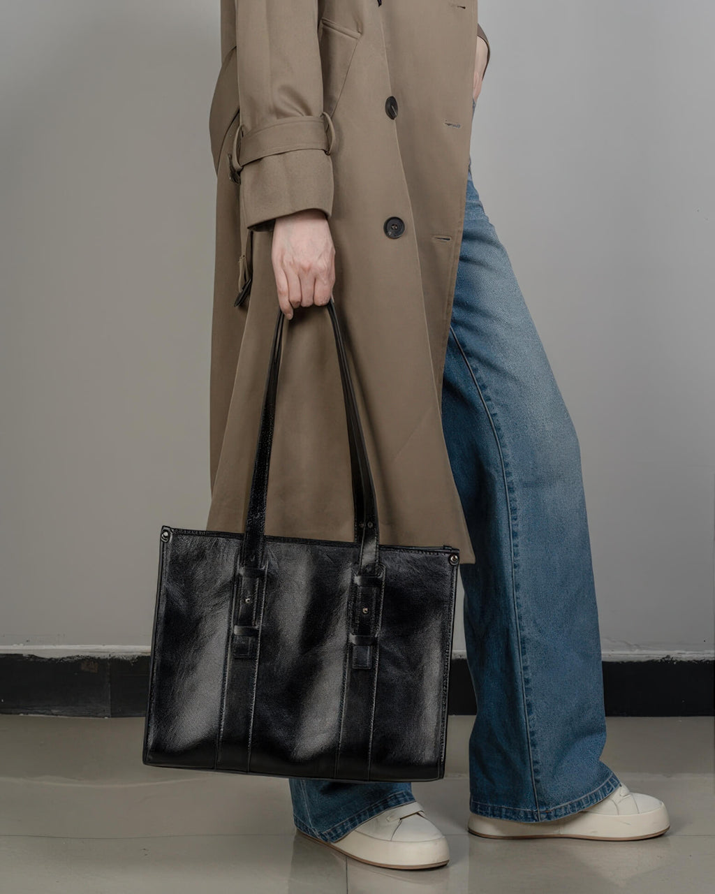 A woman in a tan trench coat holding a black leather tote bag by its top handles, styled with denim jeans.