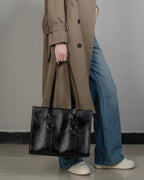 A woman in a tan trench coat holding a black leather tote bag by its top handles, styled with denim jeans.