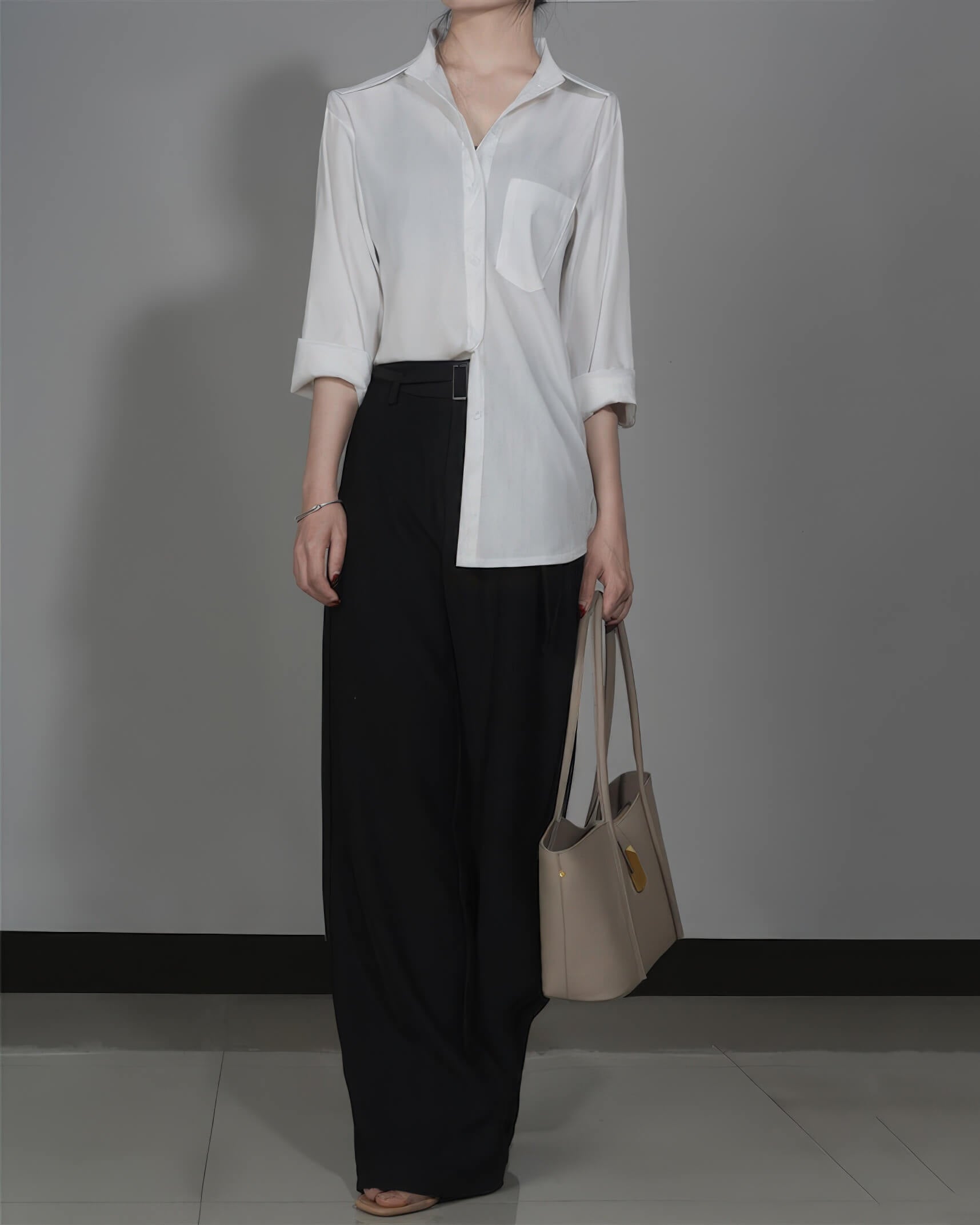 A woman in a white blouse and black trousers holding a grey leather designer tote bag by her side.