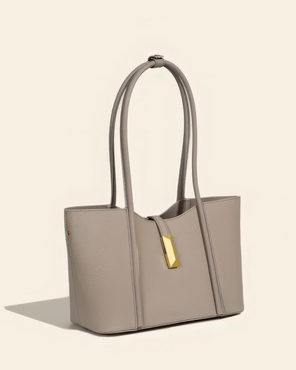 Front view of a minimalist taupe leather tote bag featuring a decorative gold vertical clasp and long shoulder straps.
