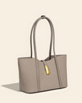 Front view of a minimalist taupe leather tote bag featuring a decorative gold vertical clasp and long shoulder straps.