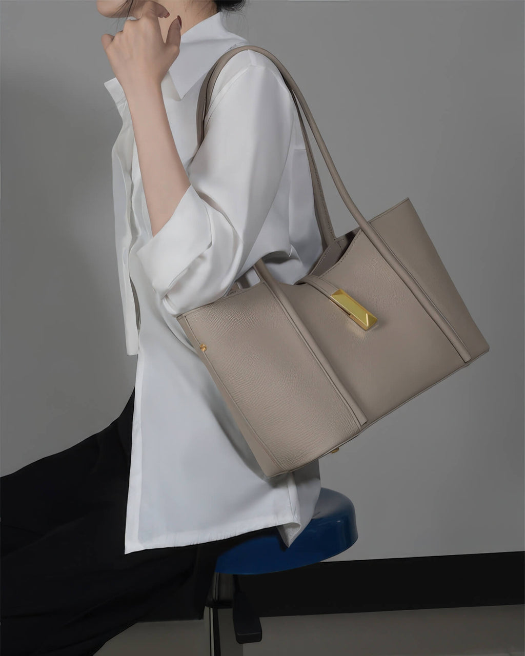 A woman sitting and carrying a high-quality grey leather handbag with gold hardware details.