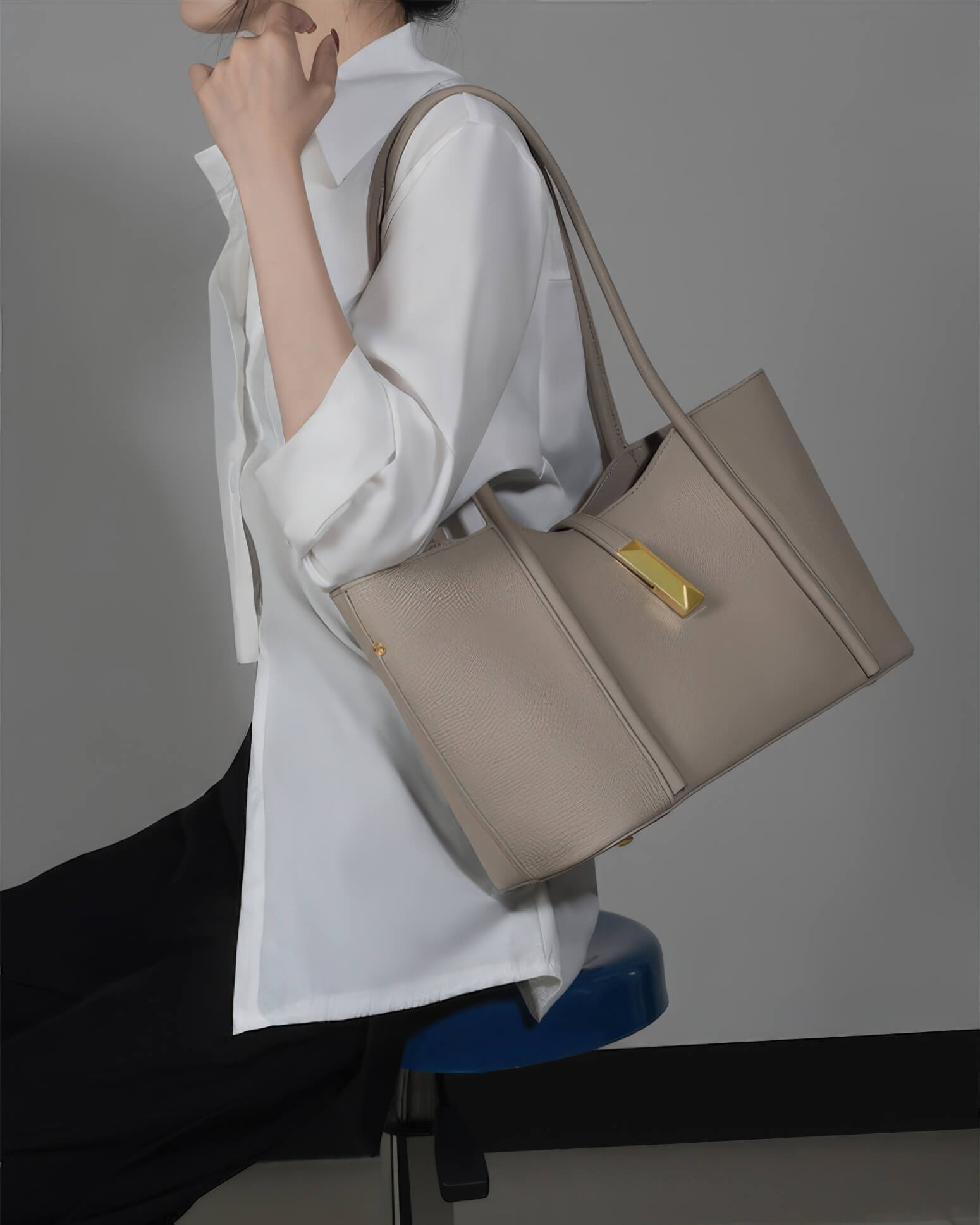 A woman sitting and carrying a high-quality grey leather handbag with gold hardware details.