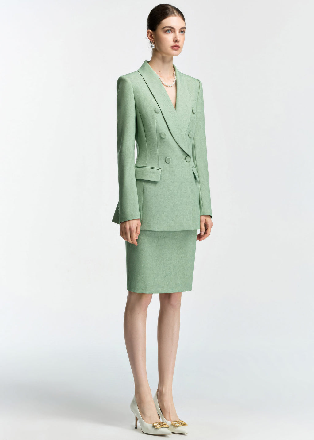 A full-body shot of a model wearing a mint green, double-breasted blazer and matching pencil skirt set. She is standing at a slight angle and wearing white pointed-toe heels with gold accents.