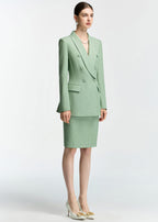 A full-body shot of a model wearing a mint green, double-breasted blazer and matching pencil skirt set. She is standing at a slight angle and wearing white pointed-toe heels with gold accents.