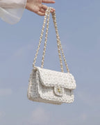 Cream woven tweed crossbody bag with braided chain strap held against clear blue sky