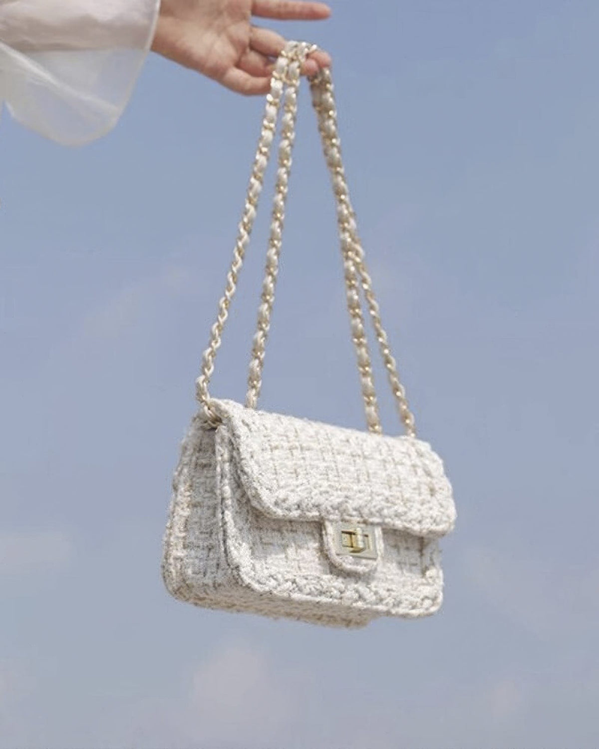 Cream woven tweed crossbody bag with braided chain strap held against clear blue sky