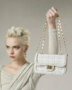 Blonde model holding up cream woven tweed crossbody bag with gold chain strap and turn-lock closure