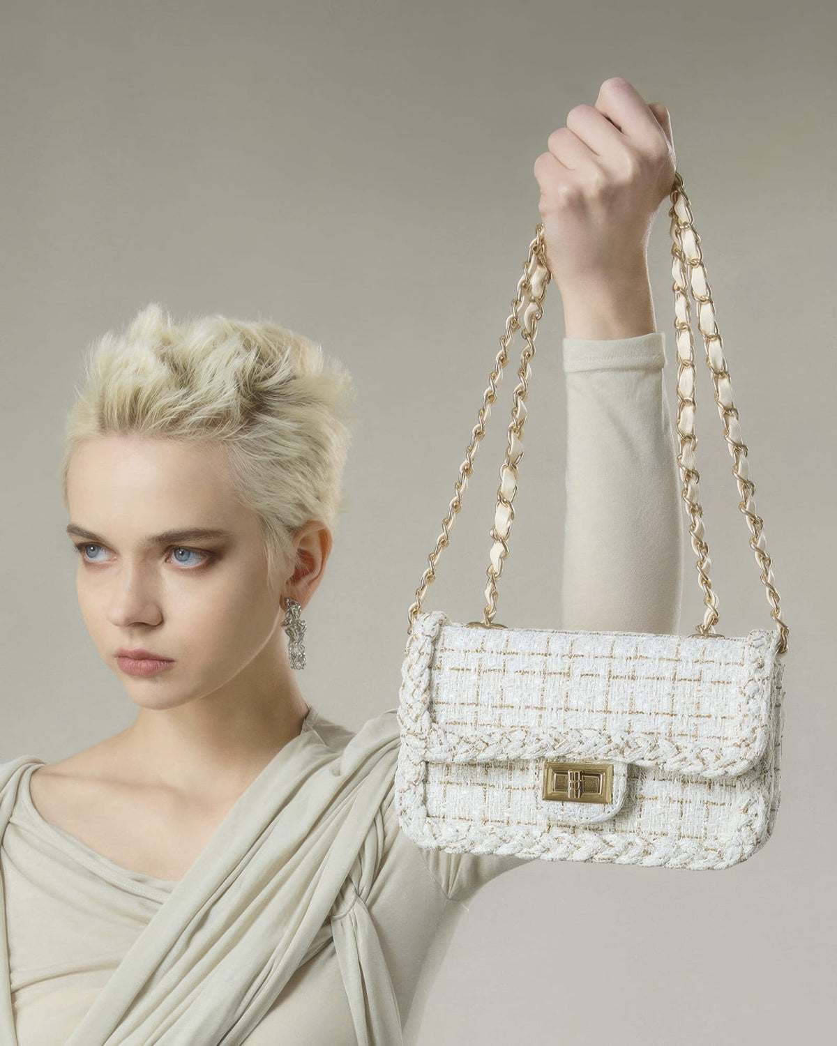 Blonde model holding up cream woven tweed crossbody bag with gold chain strap and turn-lock closure